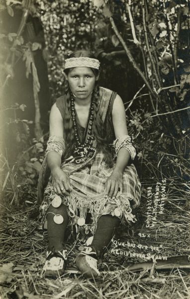 Photographic postcard of Qua-bas-er-o-qua, an Ojibwa (Chippewa) woman. She is seated on the ground in a low woven chair, and is wearing traditional clothing. Her feet are resting on a rifle. In the background are trees and foliage, and a tent on the left. Caption reads: "Qua-bas-er-o-qua, A Chippewa Squaw, Antigo, Wis."