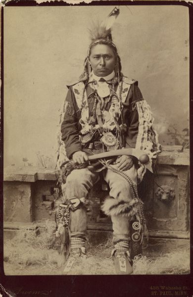 Cabinet card of Ojibwa Chief Hole-in-the Day. He is seated on a decorative stone wall in front of a painted backdrop. At his feet is dried grass. He is wearing traditional clothing and is holding a Bird Head War Club.