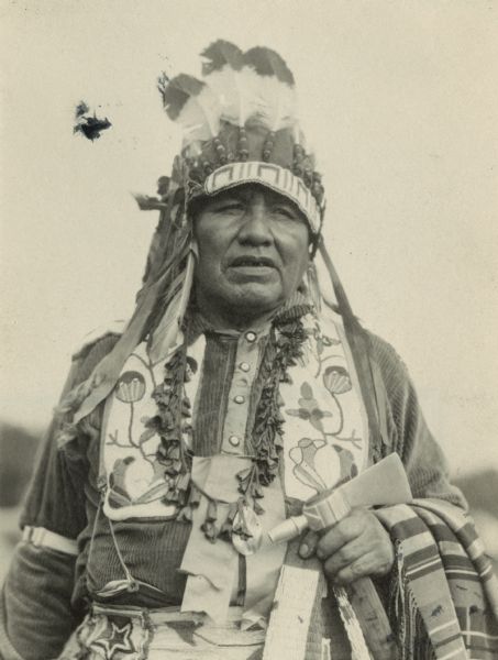 Ojibwa man dressed in traditional Native American clothing with a blanket over his arm. He is holding a tomahawk.