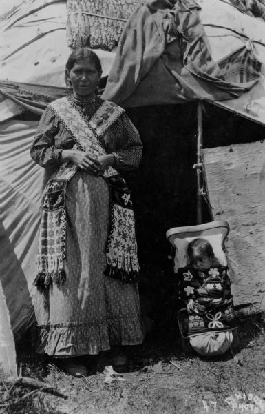 Outdoor portrait of a Chippewa (Ojibwa) woman and child posing in front of a wigwam, a waginogaan (domed-lodge). The woman is dressed in traditional Native American clothing with bandolier bags, and the baby is strapped into a backboard which is sitting on the ground leaning against the wigwam which is built of bark, cloth, saplings and rope.