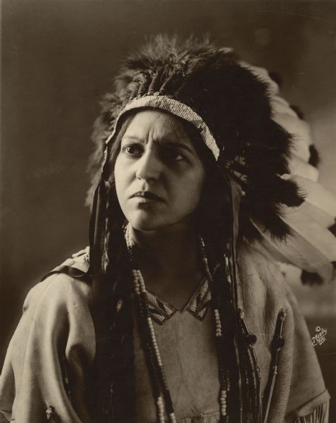 Formal quarter-length portrait of Nadonis Shawa wearing a feathered headdress and traditional Native American clothing.