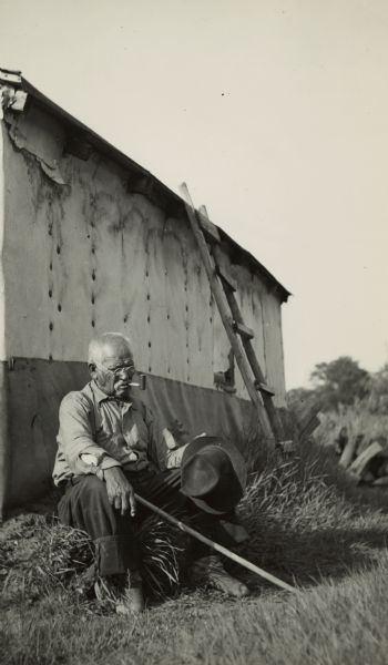 Caption on reverse reads: "An old Chippewa Indian meditates as he puffs a new-fangled pipe of peace." He is seated on a hillock of grass with a pole in one hand and his hat in the other. A deteriorated old building with a ladder is behind him.