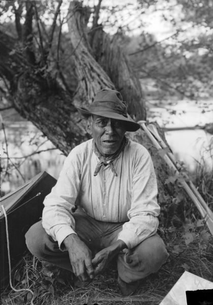 A Native American man from the Mille Lacs and Rainy Lake area in Minnesota. He is probably Chippewa (Ojibwa). Behind him are paddles leaning on a tree and the bow of a modern day canoe. In the background is a lake.