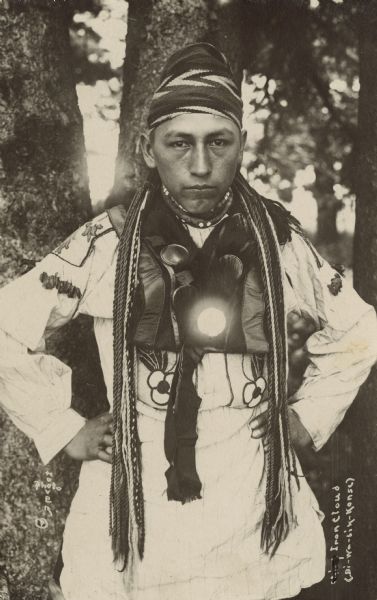 Photographic postcard of a Chippewa (Ojibwa) man, Iron Cloud, Bi-wa-bik-konse, posing in traditional Native American clothing. One of the ornaments on his chest has a reflection. In the background is a wooded area.