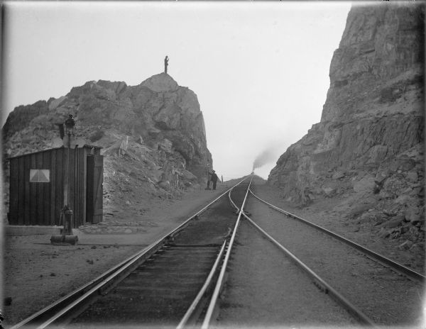 Switchman standing at the switch next to railroad tracks cut through a rock formation. Another man is standing on top of the rock formation on the left. In the distance, a man can be seen standing on a train with a plume of smoke on a railroad bridge. In the foreground two or three sets of tracks are joining together. On the foreground on the left is a small shed with a window, and another switch. The location is the Western United States. At this time Carl Peterson was writing letters home to Christine Jenson about his journey.