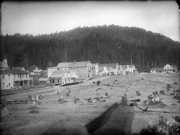 Caption on negative sleeve reads, "Mining (?) town with view of saloon, restaurant and general store." A boardwalk runs along the fronts of the buildings. In the background are forested hills, and in the foreground is a clear cut field with many stumps. At this time Carl Peterson was writing letters home to Christine Jenson about his journey.