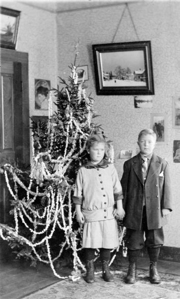 Muriel and Laurie Peterson hold hands as they pose for a photograph in front of a decorated Christmas tree. In the background are many prints on the wall and a door. On the wooden floor is a large carpet. Caption reads: "Waiting. Xmas 1913. Dec. Home-Scenes." 