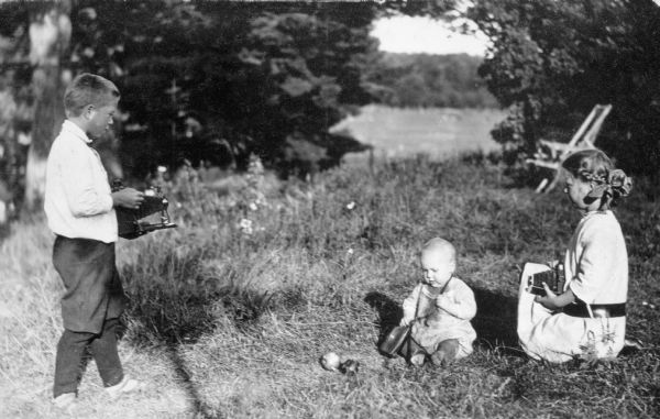 Laurie Peterson, standing, and Muriel Peterson, kneeling, pose as if taking photographs of James seated on the ground. They each hold a camera. James is tugging on a strap holding an object, hanging around his neck. In the background, the lake appears in an opening in the trees. Partially visible behind Muriel is a chair.