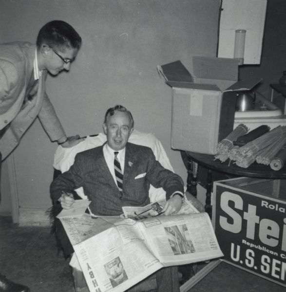 Governor Vernon Wallace Thompson, seated in a chair, is reading the newspaper and taking notes in the Republican Headquarters during his re-election campaign. Ron Emmal is looking over his shoulder. On the right is a campaign sign for Roland Stein for U.S. Senate, and a pile of supplies.