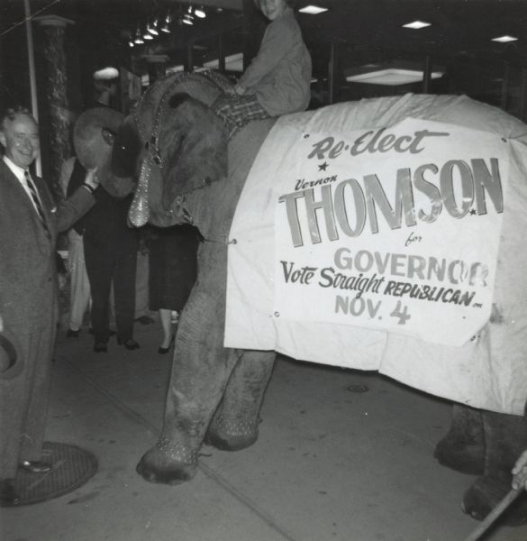 Governor Vernon Thompson (left) has his hand on the trunk of an elephant, the symbol of the Republican Party. The elephant is draped with a banner that reads: "Re-Elect Vernon Thompson for Governor, Vote Straight Republican on Nov. 4." A child is seated on the elephant's shoulders. In the background is a crowd of spectators.