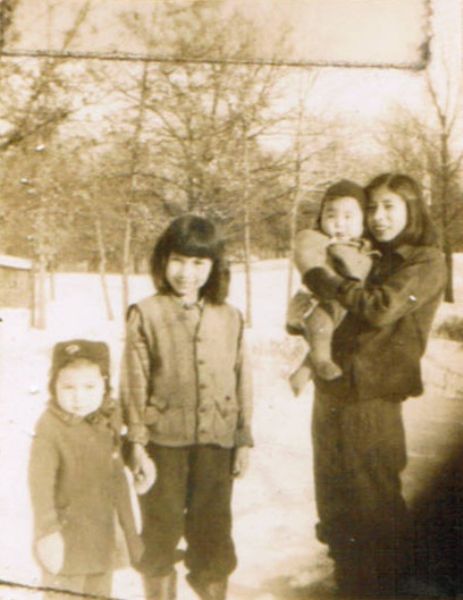 Four Native American children pose outdoors in the snow. On the right, a girl holds an infant. On the left, another girl holds a child by the hand. In the background are trees.