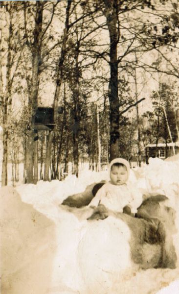 A Native American infant, nestled in a deer hide, is posed sitting up in a depression in a snowbank. In the background are trees, a dwelling and a tree house or tree stand.