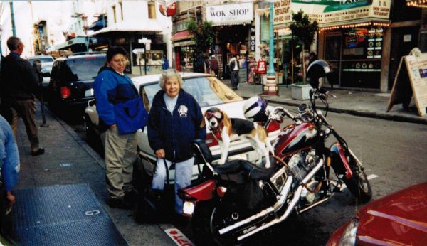 Harriet Oiyotte LaSarge poses next to a goggle wearing hound dog on a motorcycle. On the left, another Native American individual looks on. In the background is a street in a business district of an unknown town, automobiles are parked along the curb.