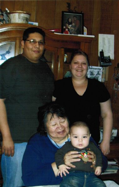 A casual, indoor family portrait featuring three generations. The grandmother (seated), holding the child on her lap, is one of the five Oiyotte sisters. In the background are a hutch, telephone, framed photographs and other household items.
