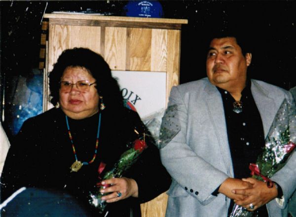 Frances Ann Oiyotte Decorah and a Native American man stand in front of a wooden podium, each holding a cellophane wrapped rose. 