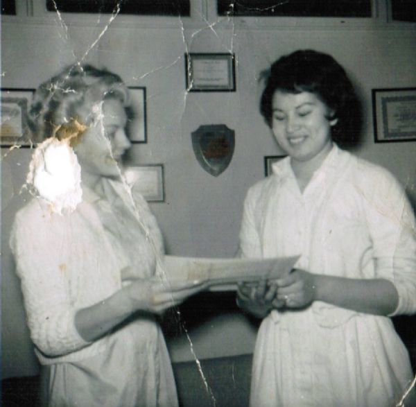 Frances "Frenchie" Ann Oiyotte Decorah receiving her beauticians license at the age of 18 from the Green Bay Beauty School. The blond woman on the left is unknown. They are wearing dresses and sweaters. The wall in the background is covered with awards and certificates.