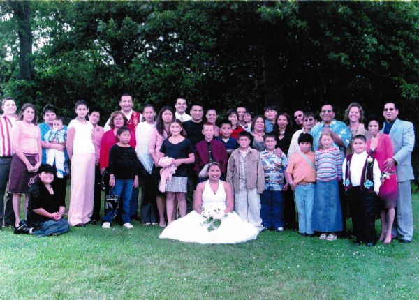 A large, outdoor, group wedding portrait of group of people posing on a lawn. [name of family to come] The bride is sitting in the center on the ground, with her skirt fanned out. She is wearing a halter style wedding dress, a feather in her headpiece, and is holding a bouquet of white flowers.
