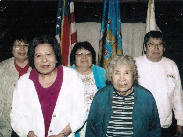 A group snapshot of the "Five Sisters." Names are, left to right: Beverly Oustigoff, Ann Marie "Gaagabiikwe" Oiyotte, Frances "Frenchie" Ann Oiyotte Decorah, Harriet Rose "Chippediikwe" Oiyotte LaSarge and Doloris M. Oiyotte Emery. The sixth (missing) sister's name is Angeline Oiyotte Johnson. In the background are American and Tribal flags.