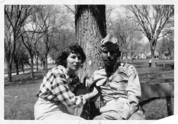 An Ojibwa man and woman are sitting together on a park bench. She is wearing a skirt and plaid blouse, and he is wearing a military uniform. Immediately behind them is the trunk of a large tree. Trees, homes and sidewalks are in the background.