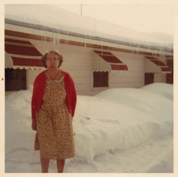 Snapshot of Mary Taylor Oiyotte standing outdoors in the snow. She is wearing a flowered dress and red sweater. Behind her is a snowbank and the windows of a white dwelling with red and white awnings. She is the mother of the "Five Sisters."