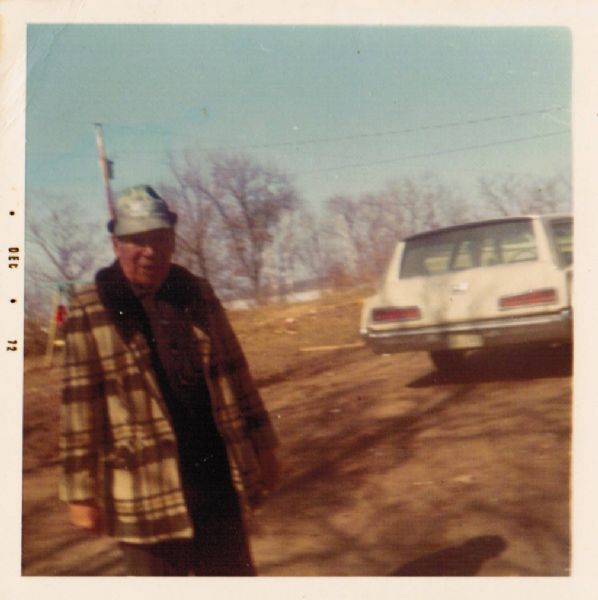 Ojibwa man standing outdoors near an automobile. The man is wearing a plaid coat, and a cap. In the background are trees and a lake.