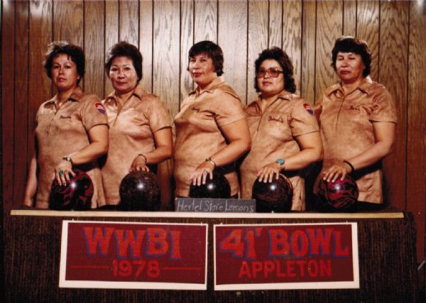 Group portrait of a female bowling team named "Hertel Store Lemons." They women are wearing matching peach-colored bowling shirts, and four of the shirts have names embroidered on the upper left side. Bowler's names (l to r): Marian Reynolds, Ann Oiyotte, Beverly Oustigoff, Frenchie Decorah and Doris Emery. The tournament is named the "WWBI 1978" at the "41' Bowl Appleton."