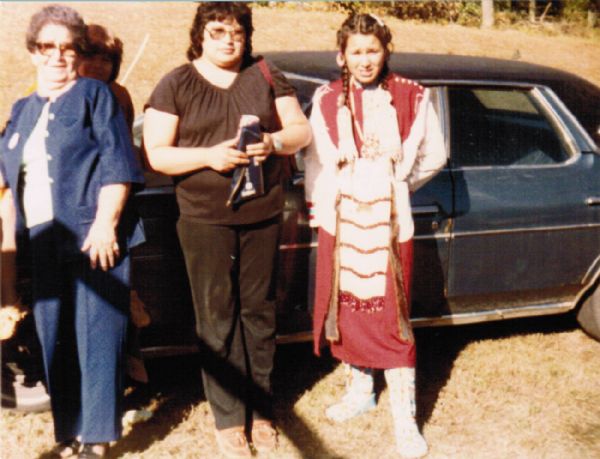 Three Ojibwa women are standing in front of a car. A young person is sitting on the hoodoo the car, and is peeking over the older woman's shoulder. The young woman on the right is dressed in Native American ceremonial regalia. Names, standing, (l to r): Ruby Taylor, Frances Decorah and her daughter Kimberly Phernetton.