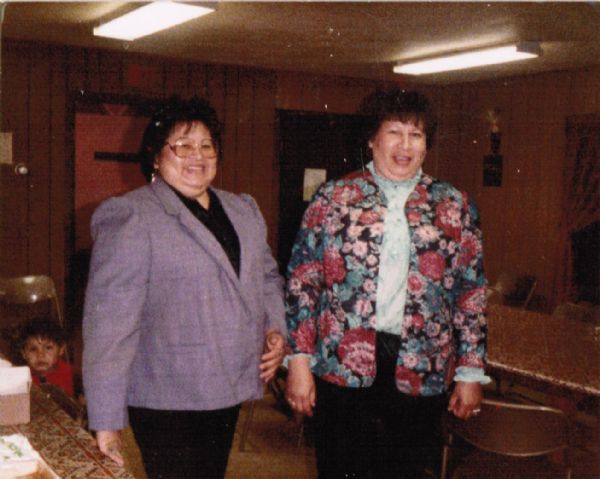 Two of the "Five Oiyotte Sisters" pose together at a daycare. On the left is a child seated in a chair. The sisters attended early childhood classes in Rice lake, then obtained a grant to start a daycare.
