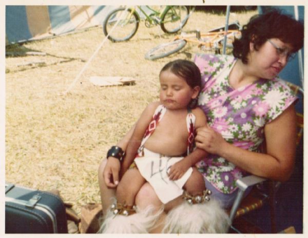Frances Decorah and her son Patrick at a Pow-wow in South Dakota. She is seated in a lawn chair and Patrick is napping on her lap. Frances is wearing street clothes and Patrick is wearing Native American ceremonial dancing regalia with bells and fur leggings. He has a number on his outfit. On the left is a suitcase, in the background are two tents and two bicycles.