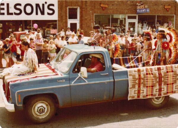 Stuart (husband of Frances Decorah) driving a truck in a street parade. The truck is draped with blankets displaying a Native American motif. A woman in traditional Native American garb is perched on the hood and four youth are standing in the truck bed, similarly dressed. A crowd is watching from the curb on the far side and storefronts with signs for "Nelson's Shoes" fill the background. 