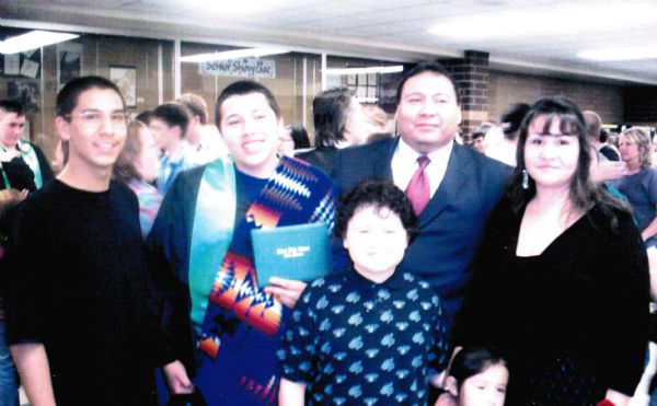 Group portrait of six people at a high school graduation. The graduate is wearing a blanket with a Native American motif draped over one of his shoulders, and is holding his diploma. In the background is a crowd of other families.