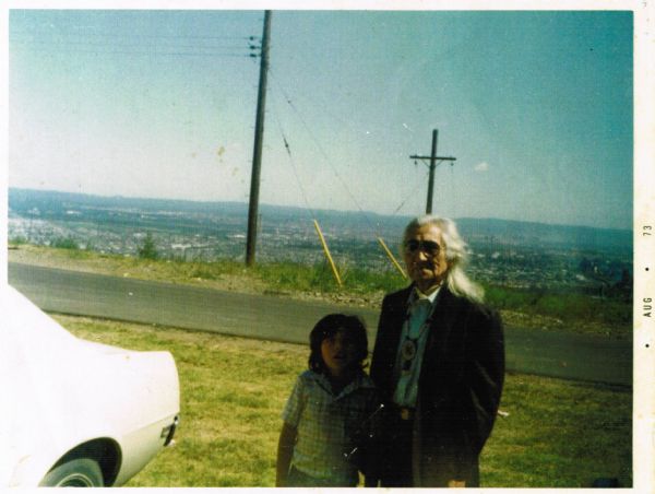 A man and youth posing together outdoors next to the back of a white automobile. The man is wearing a necklace containing ornamental beading. In the background is a blacktop road, power poles and a distant view of a community with hills on the horizon.