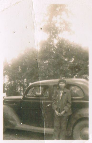 A Native American woman is posing outdoors while leaning against an automobile.