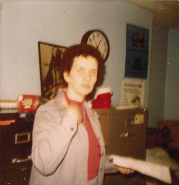 An unknown woman posing in an office with papers in her hand. In the background on the wall is a print of a Native American and a clock above filing cabinets.