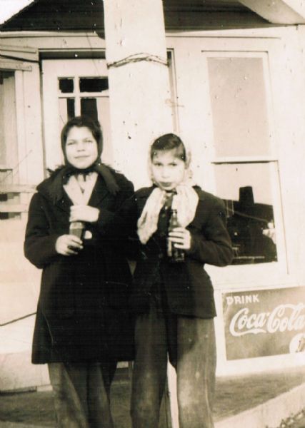 Two young women posing standing outdoors holding soft drinks in bottles. On the right is a "Drink Coca-Cola" sign. They are wearing coats and scarves. 