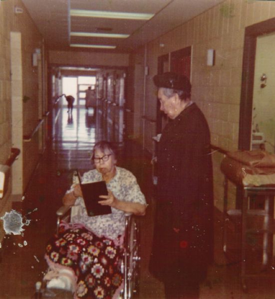 Two women in a hospital corridor. The woman on the left is sitting in a wheelchair reading a book, with one of her feet in a "boot." She is wearing a hospital gown and has a "granny square" afghan on her lap. The woman on the right is standing and wearing a coat and hat.