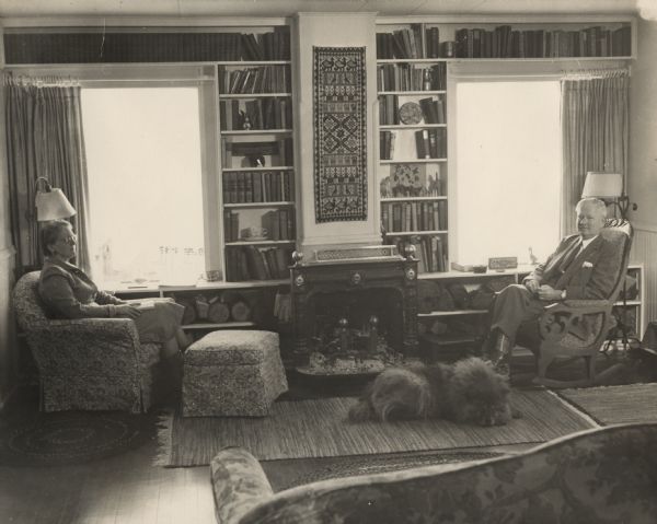 Phillip and Isabel Bacon La Follette sitting in comfortable chairs in their living room at home. A large dog, perhaps a Chow, is lying on the rug in front of a wood-burning stove between two windows. Built-in bookshelves surround the sides and tops of the windows, and firewood is stacked on the shelves below the windows on either side of the stove. A woven piece of artwork is hanging between the windows. In the foreground is the back of a sofa.