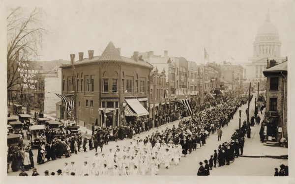 Photographic postcard of an elevated view of a patriotic parade marching from the Capital towards lower State Street. Spectators are lined up and watching from along the sidewalks. Shows the even side of the 200 block of State Street. Cars are parked on W. Johnson Street. Handwritten on the left side: "The Kamera Kraft Shop #6."