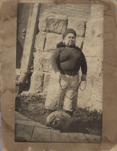A man is posing with a badger on a leash by the Armory on the University of Wisconsin-Madison campus. He is wearing thickly quilted, light-colored pants, a belt, a dark sweatshirt and a knit cap with a tassel.