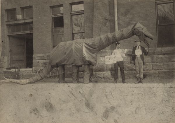 Two men are standing on either side of a large costume of a six-legged dinosaur in front of the Armory on the campus of the University of Wisconsin-Madison.  Approximately 33 feet long, the costume of a brontosaurus (an unprecedented fossil discovery around this time in the United States) is maneuvered by three people from the inside. It was created of some sort of fabric over a framework of wire. One man standing on the left and wearing a sweater, smoking a cigar, is holding the tip of the tail and a sign that reads: "WHAT IS IT?" The other man to the right wearing a suit and military hat is in front of the neck, holding a two handled cup (a trophy) in one hand and a furled umbrella in the other. They are standing In the foreground is a wood plank sidewalk and a lawn. In the Armory, guns are visible on a rack just inside the door. To the left is a wooden chair and a Pierce bicycle; on the right a ladder.
