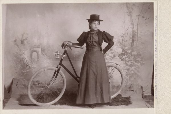 Full-length studio portrait of Pearl Miner standing in front of a painted backdrop holding a bicycle. She is wearing a dark dress and hat, and is standing on a drop cloth with a piece of fake grass on it.
