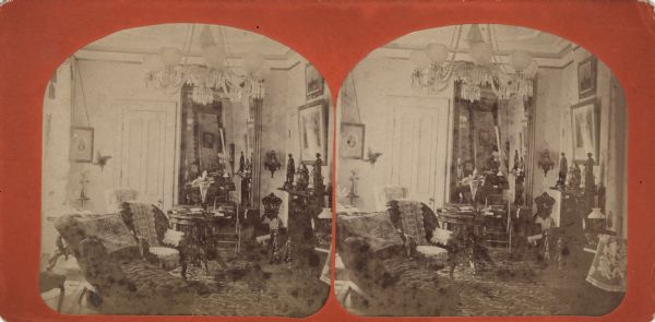 Stereograph of a room in Ole Bull's Madison residence, where he lived after marrying Sara Chapman Thorp in 1870. The room is furnished with chairs, a table, a large chandelier and a mirror, and is richly decorated with sculptures and framed images on the walls.