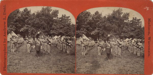 Stereograph of a music band in uniform, standing in formation on a lawn. The band major is standing in front with his baton, ornate uniform and tall hat. In the background are tents and trees. Text on reverse, "The Beauties of the City of Madison and Vicinity. Music Band."