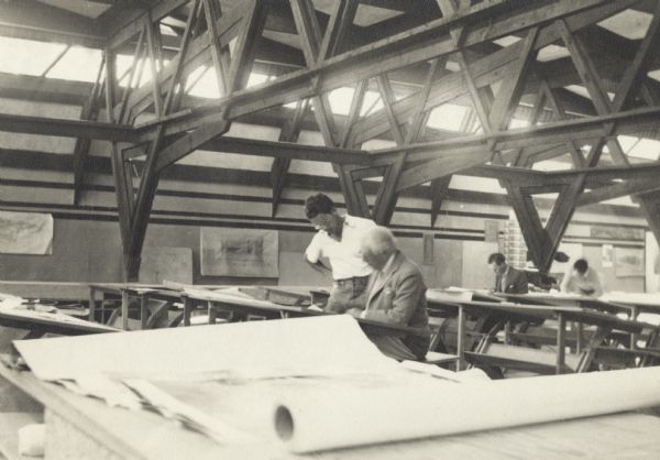 Bennie Dombar standing next to Frank Lloyd Wright, who is working at a drafting table. Trusses are overhead.