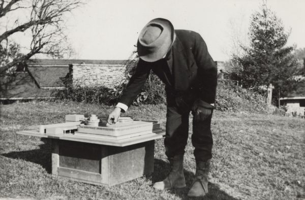 Frank Lloyd Wright standing outdoors and leaning over an architectural model.
