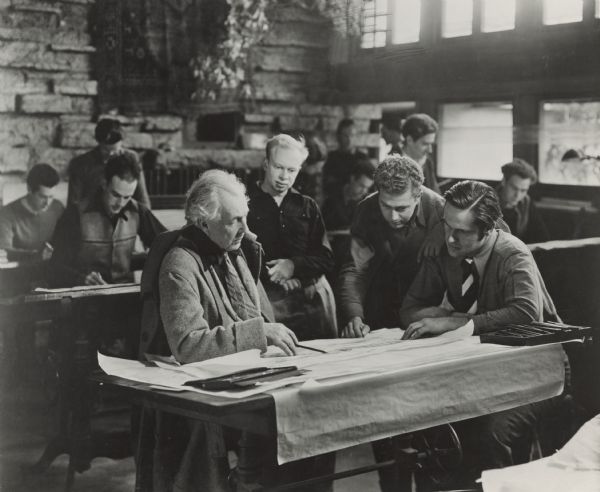 Frank Lloyd Wright is sitting at a drafting table surrounded by apprentices.