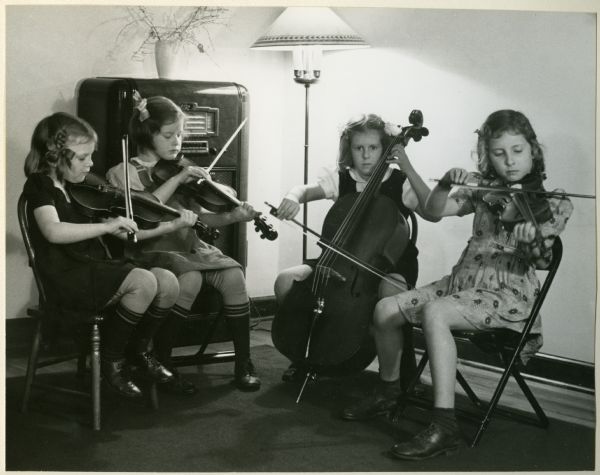 Four girls sitting and playing in a Madison youth string quartet. In the background is a lamp and radio. Names, left to right: Jane Parking, Janet Hart, Nancy Hart, and Polly Richardson.
