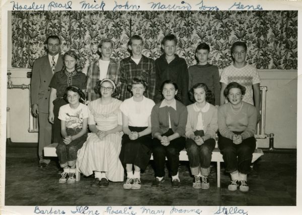 Badger School 8th grade class portrait. The front row is sitting on a bench and the back row is standing. Some of the girls are wearing saddle shoes. In the background is patterned drapery above a hot water register. Names, front row, from left: Barbara Normington, Irene Pfister, Rosalie Kaether, Mary Graves, Joanne Schuepbach, Stella Middleton. Back row: Mr. Hoesly, Pearl, Clifford Olson, John Olson, Marvin Bjornstad, Tom P., Eugene Bjornstad.