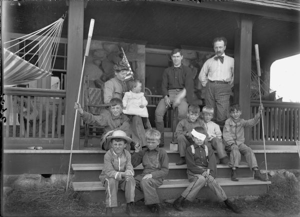 Group portrait of children and adults on porch steps. On the left a woman is sitting in a chair with an infant on her lap and an American Flag behind her. On the right are two men, one sitting on the corner of a table and one standing. Young boys are sitting on the steps. One boy has a bandage around his head and is holding a fired bottle rocket tube. Two boys are holding large unfired bottle rockets on long sticks.