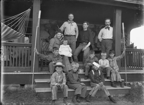 Group portrait on the porch steps. On the left a woman is sitting in a chair with an infant on her lap and an American Flag behind her. On the right are three men, one sitting on the corner of a table and two standing. Young boys are sitting on the steps. One boy has a bandage around his head and is holding a fired bottle rocket tube. Two boys are holding large unfired bottle rockets on long sticks.